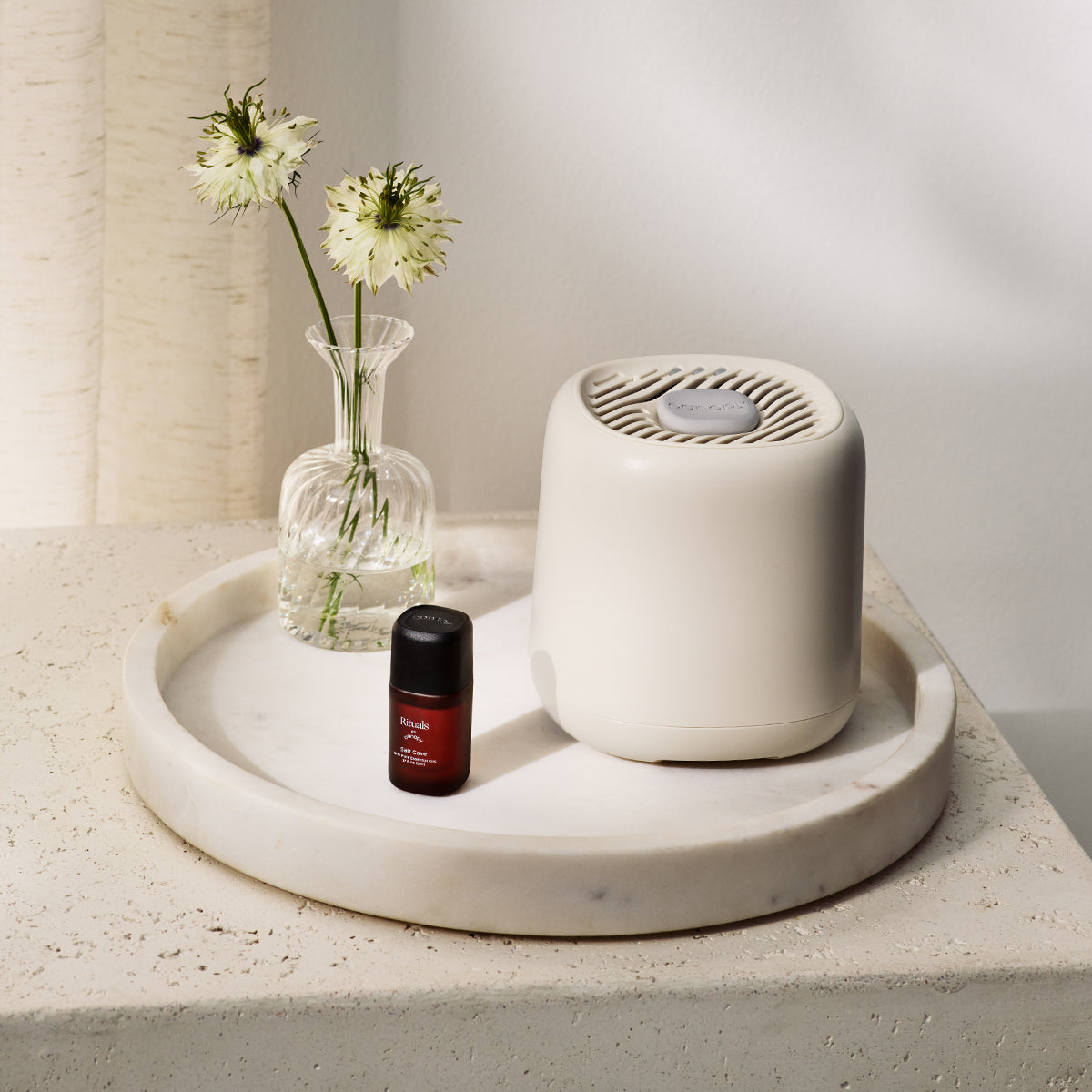 A small, white Aroma Diffuser sits elegantly on a round marble tray next to a glass vase with delicate white flowers and a bottle of essential oil, enhancing the beauty of the light-colored stone surface. The serene scene against a beige curtain and white wall reflects a touch of skincare luxury.