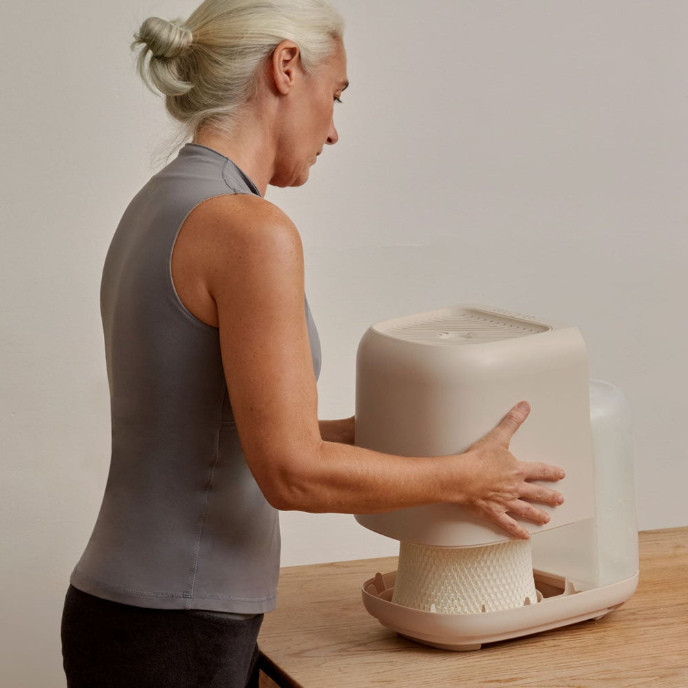 Side view of Large Room Humidifier Device Filter & Aroma being handled on wooden surface, showcasing its modern beige design with textured base and transparent water container. Premium home comfort device demonstrating easy filter access and maintenance by Canopy.