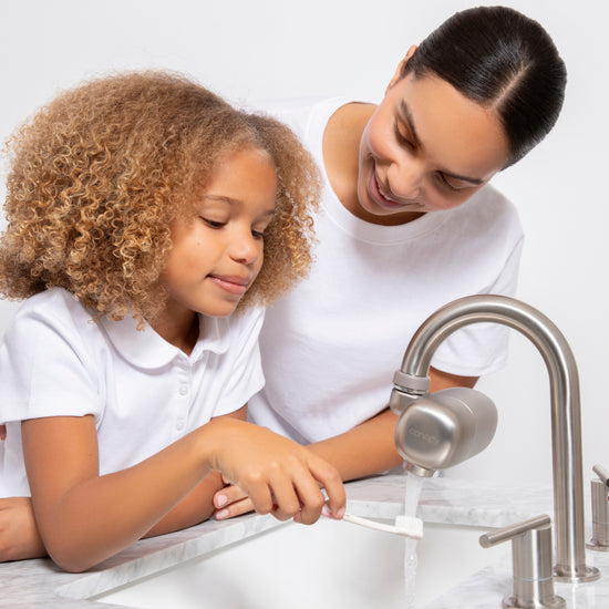 Mom is gently holding daughter by the sink, both wearing white, while the daughter is wetting her tooth brush using the Faucet Filter in Brushed Nickel