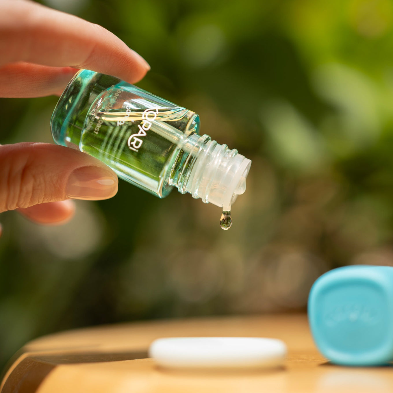 Kopari Aroma Kit glass bottle with clear essential oil dropper releasing a single drop, photographed against a soft-focus natural green background, showcasing the product's premium quality and purity. Turquoise cap visible in background on wooden surface. By Canopy.