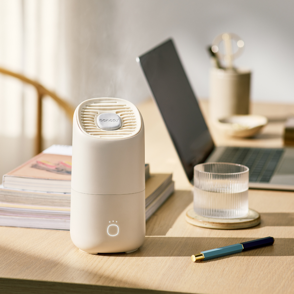 A compact Portable Humidifier sits on a wooden desk next to a closed laptop, books, and a glass of water. A pen lies nearby amidst the beauty of a softly lit room, creating a calm workspace atmosphere perfect for skincare routines.