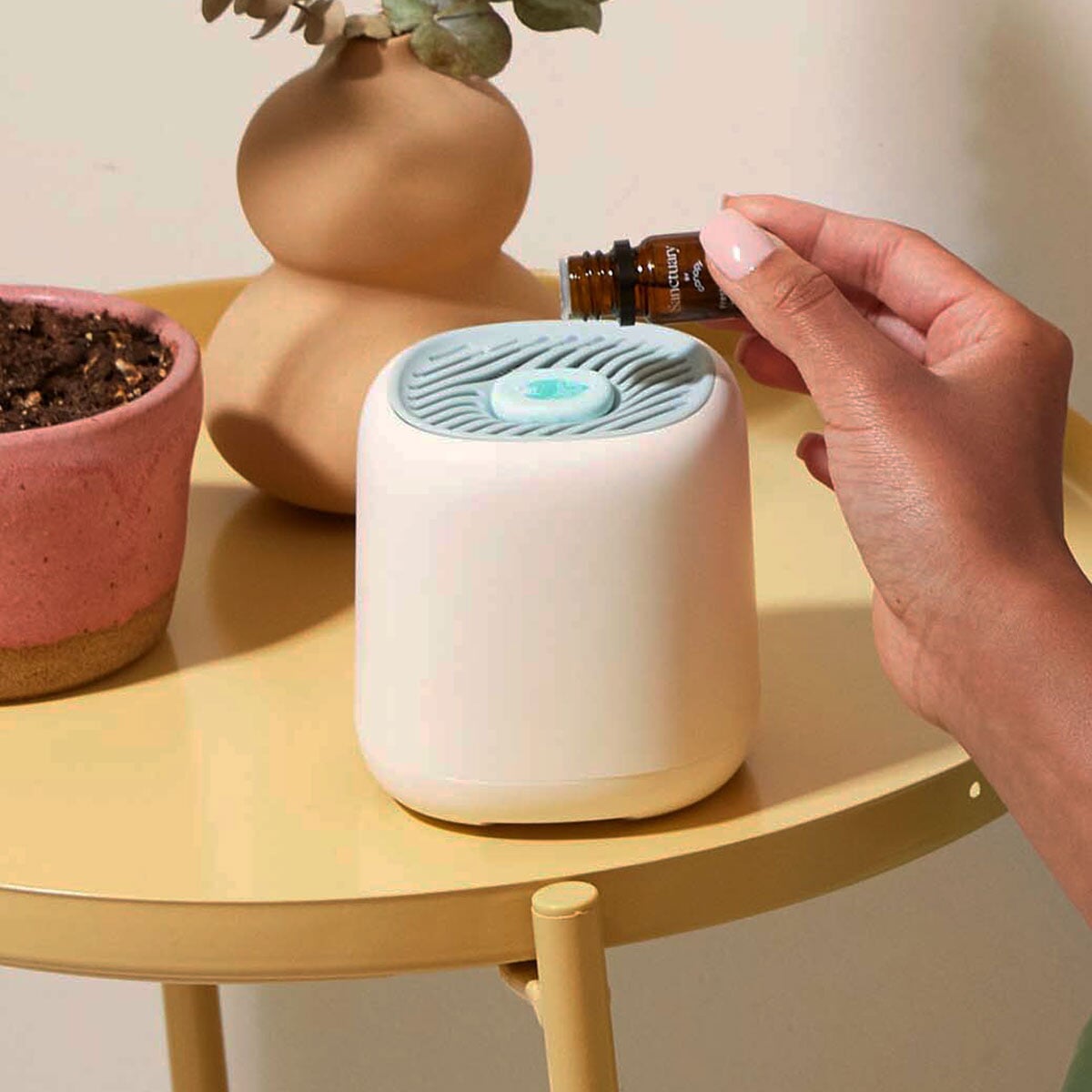 A hand adding essential oil drops to a sleek white Diffusion Well & Aroma Kit on a modern gold table, next to terracotta planters with greenery. Features a blue-gray ventilated top with glowing center. By Canopy.