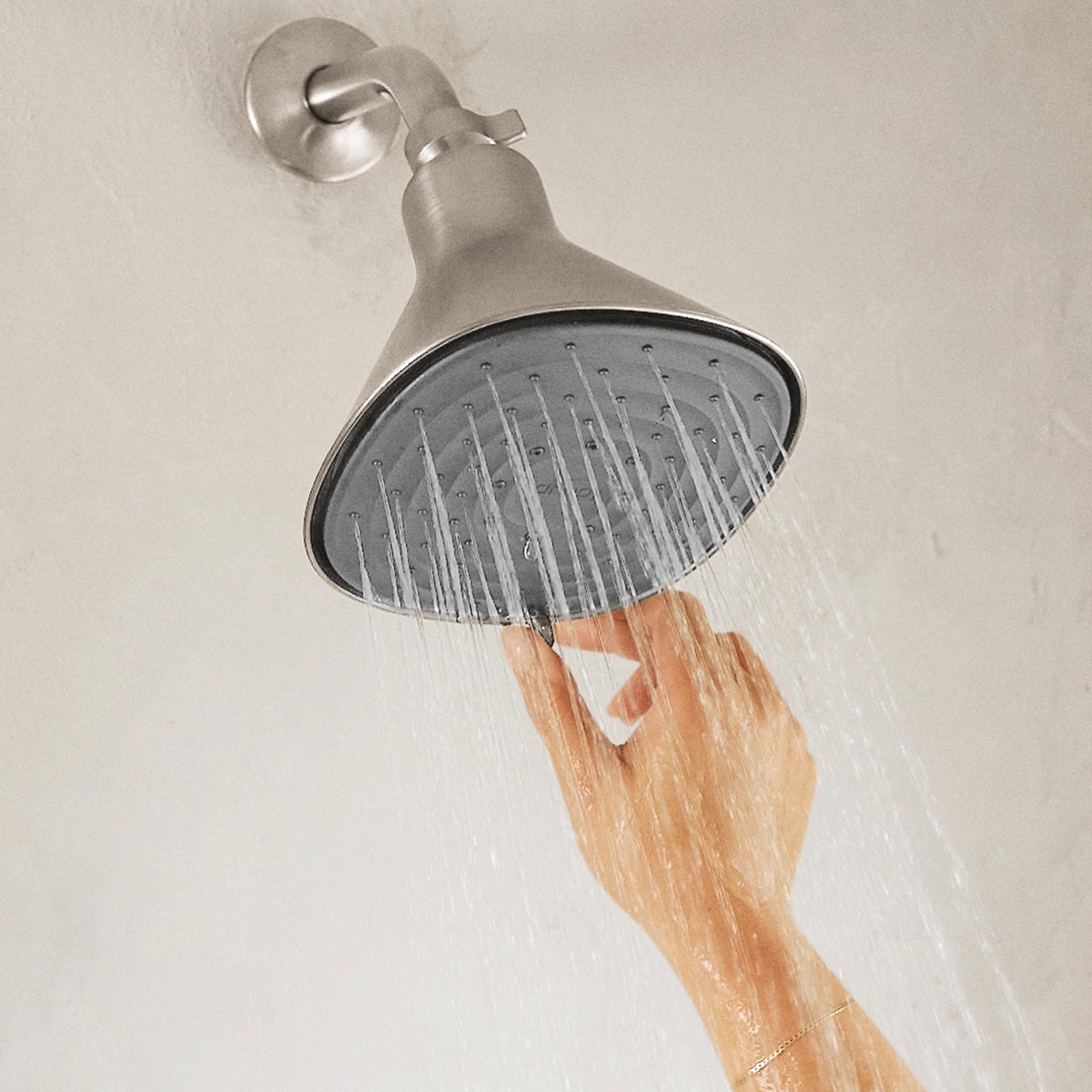 A hand reaches up to adjust the angle of a large, Brushed Nicked Filtered Showerhead, enhancing your beauty routine with a cascade of multiple water streams. The diffuse, light-colored wall in the background gently complements your relaxing skincare sanctuary.