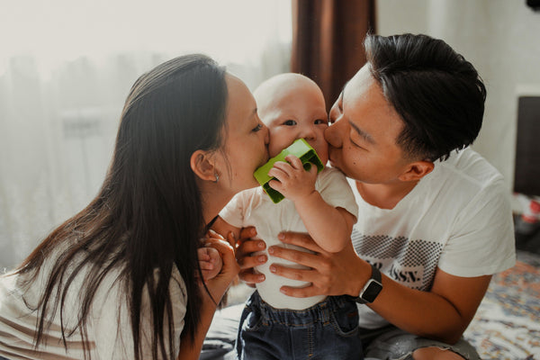 Parents holding and kissing baby, who is chewing on a green toy