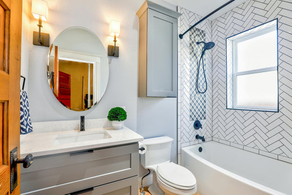 View of small bathroom with white toilet, bathtub, and sink; gray cabinets, a round mirror, light gray walls , and a herringbone patterned tile tub surround