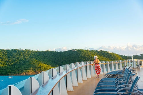 Woman in red flowered sarong standing on cruise ship deck looking at lush green mountains on a sunny day