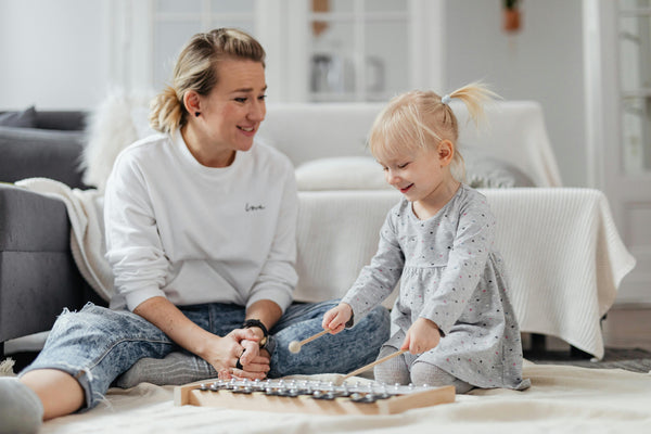 Mother and toddler daughter playing with xylophone on living room floor in front of gray sofa draped with white blanket