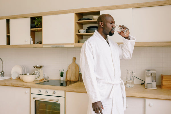 Man in white bathrobe drinking glass of water in sunny kitchen