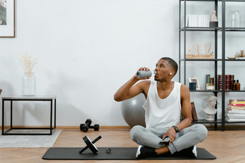 Young man sitting in modern apartment with fitness mat, ab roller device, and water bottle; hand weights and workout ball are in the background