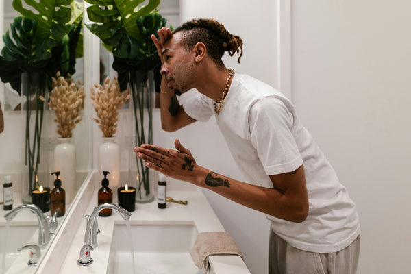 Young man washing face at the sink of a modern white bathroom with plants in the background