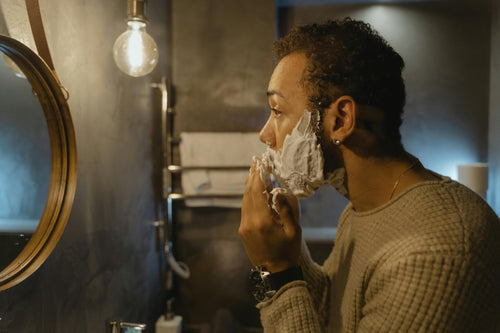Young man with shaving cream on face in front of bathroom mirror