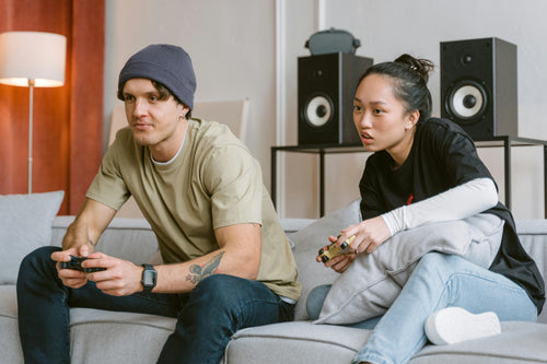Young man and woman playing games on living room sofa with large speakers in the background