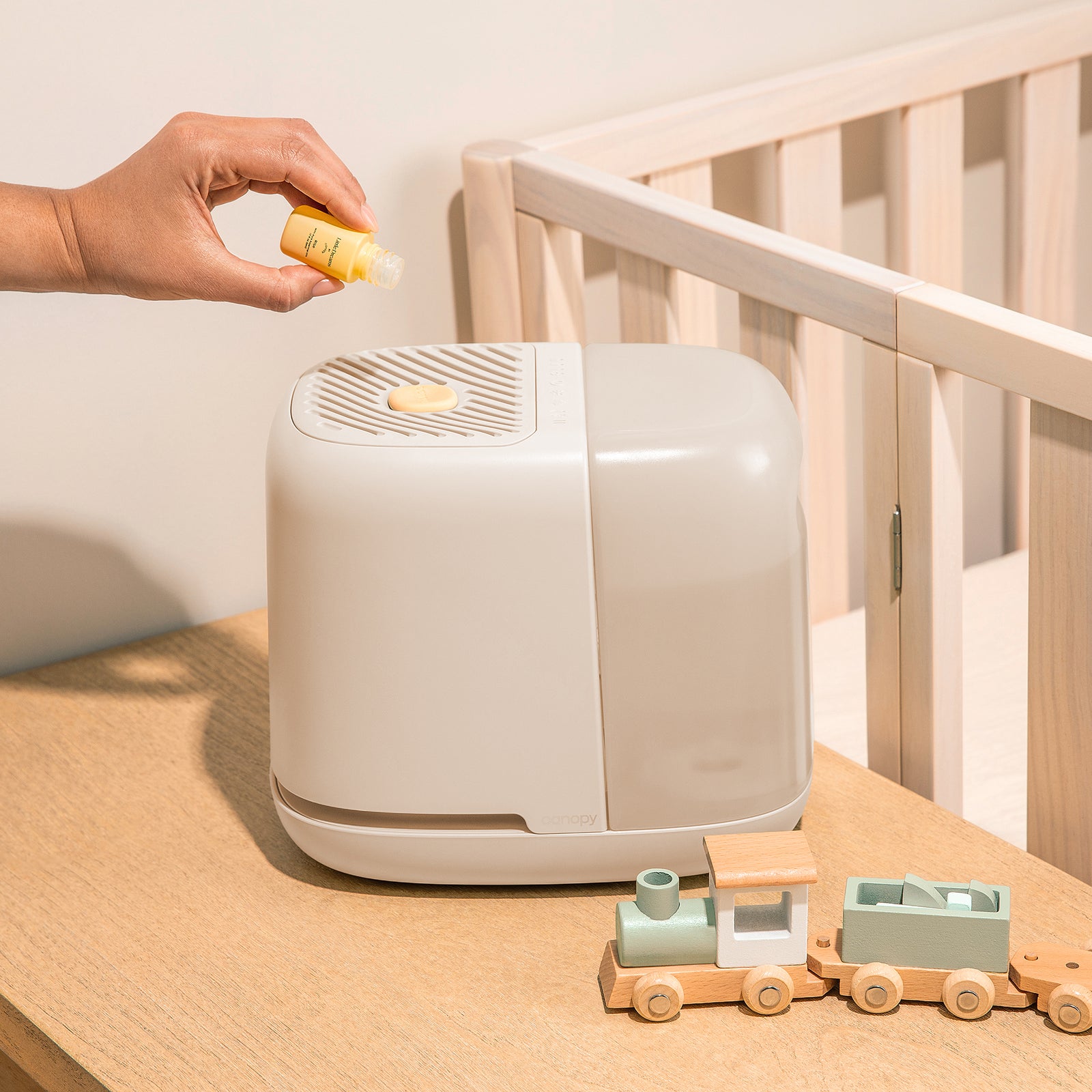 Nursery Humidifier 2.0 in Cream on a table near a crib while a hand is dripping Little Dreams Aroma Oil on to the Aroma Stone. A small green and being wooden train toy is pictured nearby.