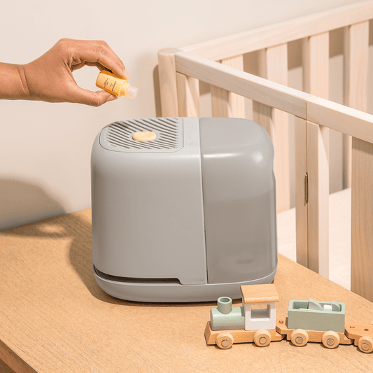 Hand adding essential oil to Little Dreams Aroma Kit by Canopy, displayed next to wooden toy train on nursery dresser beside crib, featuring sleek grey humidifier design with built-in aromatherapy diffuser by Canopy