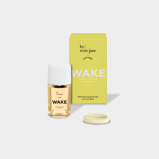 Wake Aroma Oil in elegant glass bottle with golden-hued essential oil blend, displayed alongside yellow packaging and white aromatherapy tablet. Natural energizing wellness product for morning routines. by rosie jane - Canopy