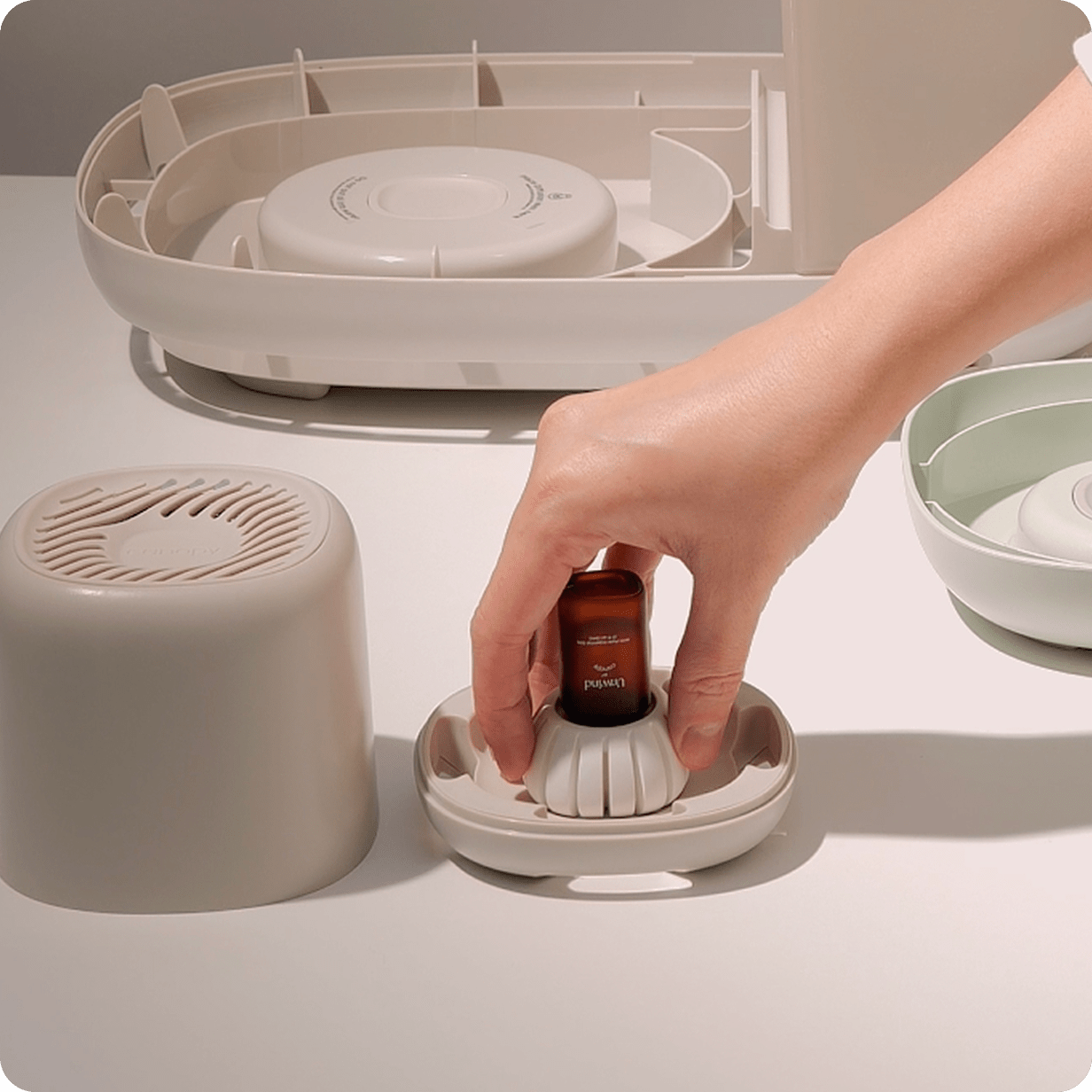 Hand placing an essential oil bottle into a white ceramic Diffusion Well 3-Pack, shown on a dark wooden table next to a terracotta plant pot, demonstrating the simple setup process for long-lasting aromatherapy by Canopy