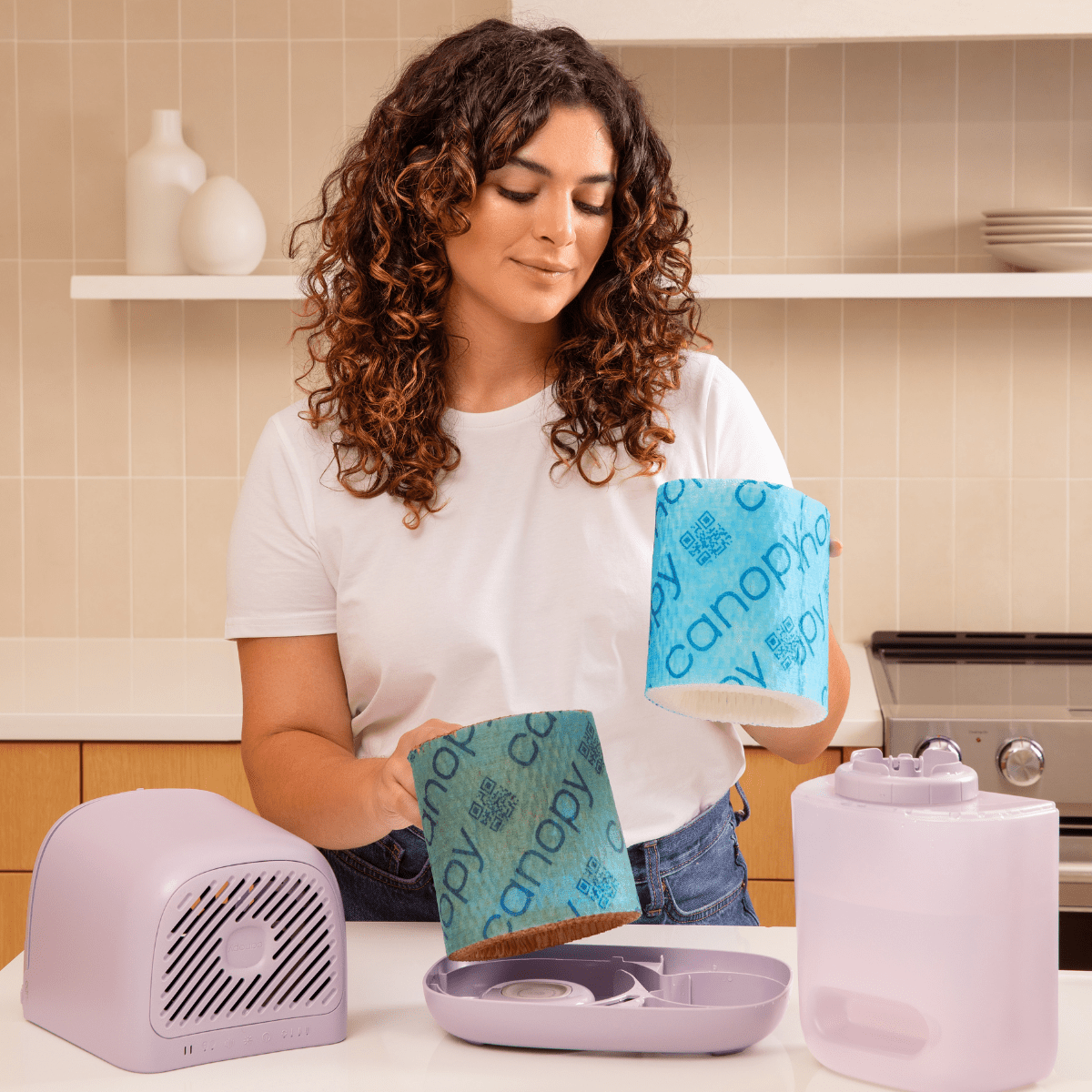 A person wearing a white t-shirt holding two replacement filters while demonstrating how to change the Bedside Humidifier Filter 2.0, with the compact gray humidifier unit and clear water tank displayed on a wooden kitchen counter with cream-tiled backsplash and floating shelves